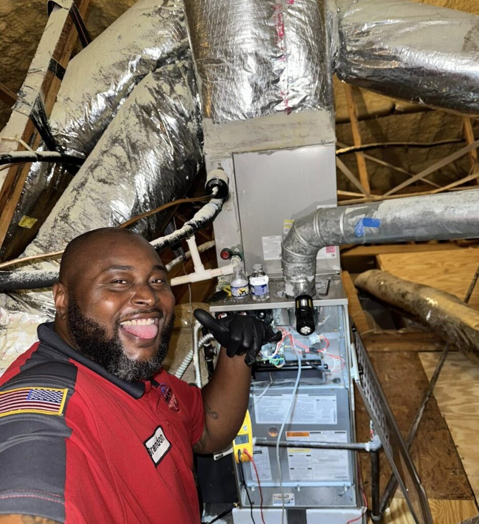 Technician in red uniform smiling and giving a thumbs up next to HVAC system in an attic, showcasing B Mays Heating & Air Conditioning's commitment to reliable service and customer satisfaction.