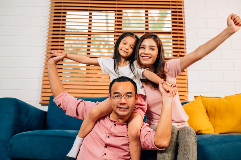 Family playing together in a cozy living room, with a mother and daughter smiling joyfully while the father lifts the daughter on his shoulders, emphasizing warmth and togetherness in a comfortable home setting.