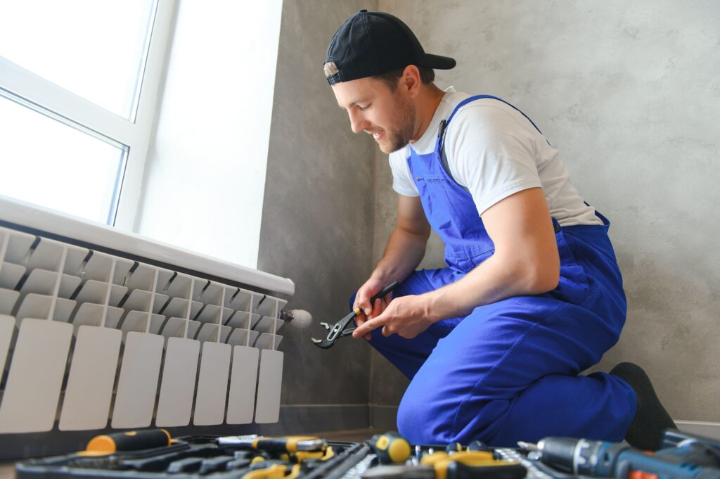 Technician in blue overalls repairing a heating radiator, surrounded by tools, in a well-lit indoor setting, emphasizing heating system maintenance services.