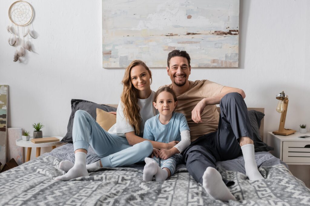 Family of three smiling together on a bed in a cozy room, representing comfort and togetherness, relevant to B Mays Heating & Air Conditioning's focus on creating a comfortable home environment.