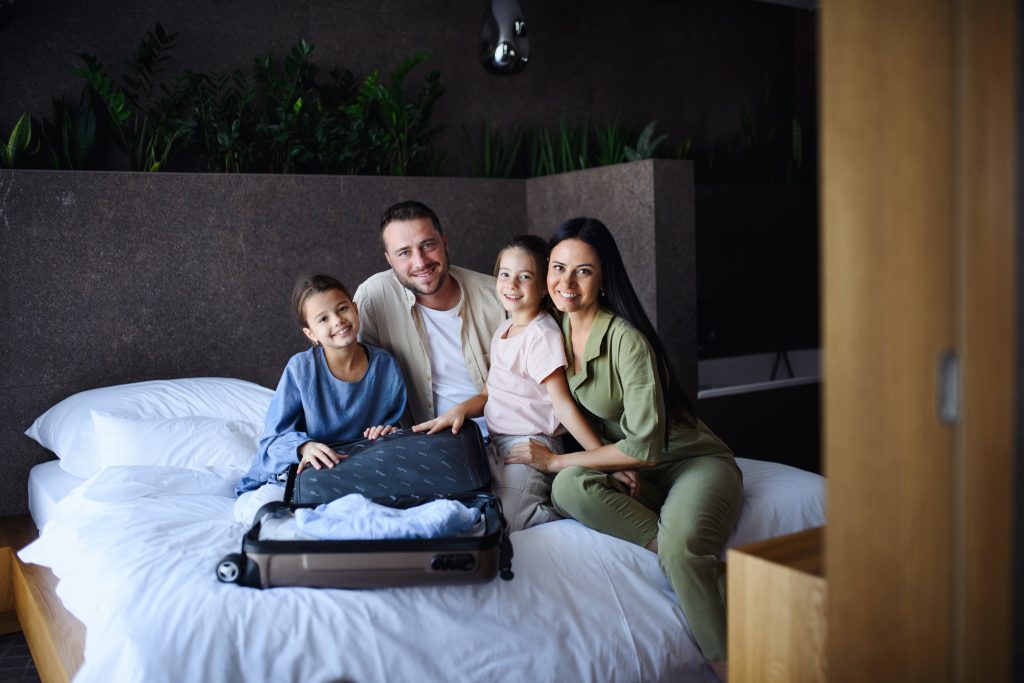 Happy young family with two children sitting on a bed with an open suitcase, preparing for a trip, in a modern, cozy room.