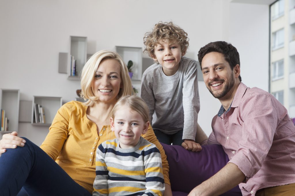 Happy family of four sitting together in a cozy living room, smiling and enjoying time at home, representing comfort and family life.