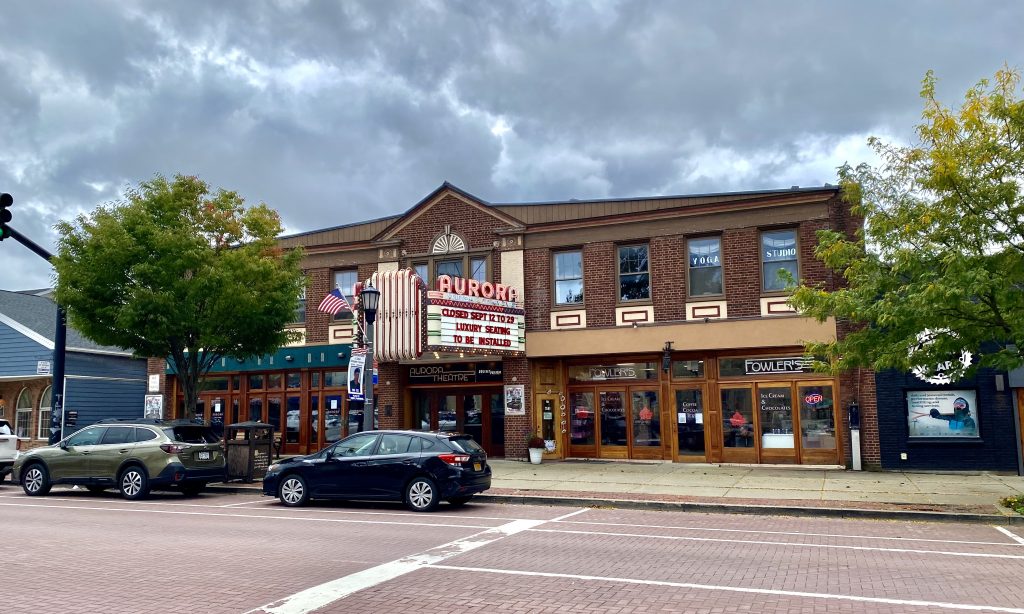 Aurora Theatre in East Aurora, New York, featuring a marquee, American flag, and adjacent businesses, with trees and cars in the foreground.